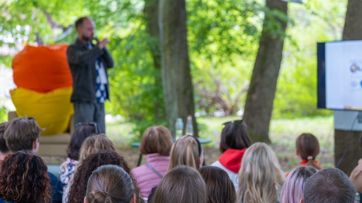 Audience in the foreground with a speaker in the background, all surrounded by trees
