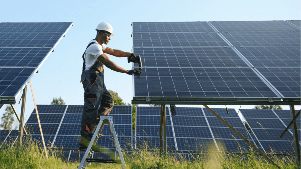A person in a white hard hat stands between two solar panels, working on the one to the right