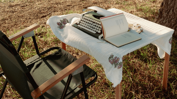 A green folding chair sits to the left of a table with a white cloth on it; on top of the table is a green typewriter and open book; behind this the ground is covered with yellow grass