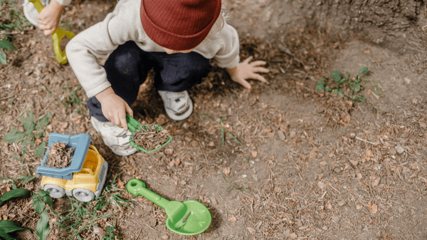 A child digs in the dirt with a green plastic shovel, below them is a plastic truck and another shovel