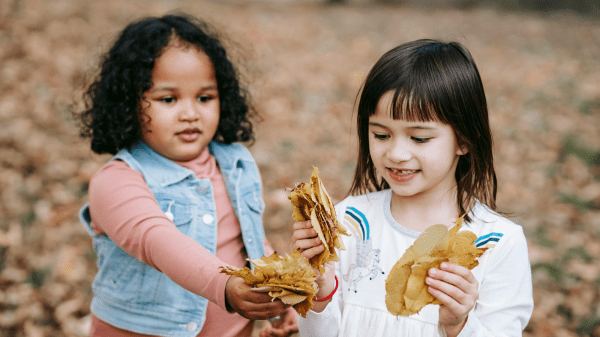 Two children hold clumps of brownish orange leaves, behind them is a blurred background where leaves can be seen on the ground
