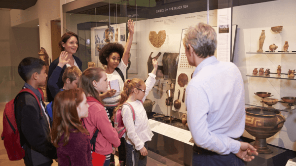 A group of five children stand in front of an adult, three raising their hands; to the right is an adult facing them; behind all of them is a display of fossils