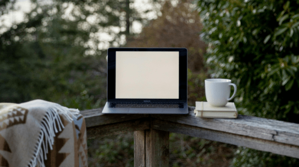 An open laptop sits on a patio fence between a blanket and a coffee mug; behind it is a forest of trees