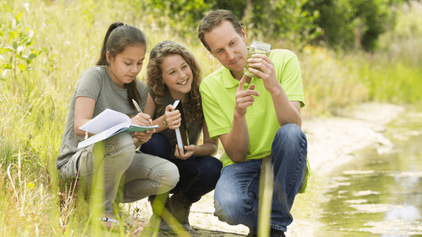 Three people squat near a creek to their right; the person on the very right holds a jar of water that they are pointing to, the person on the very left is writing in a notebook