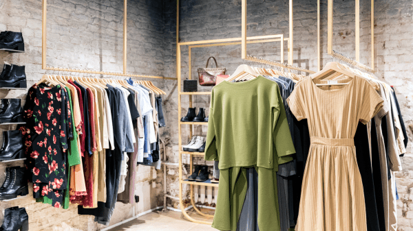 A brick-walled room contains a shelf of shoes, rack of clothes, a shelf of shoes and a handbag, and an additional rack of clothes