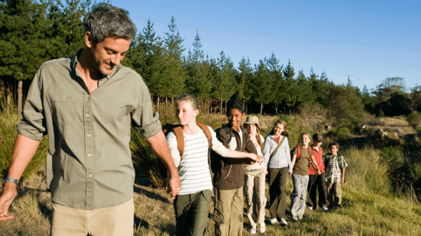 A teacher leads a group of children single file along a trail near a forest. The children follow closely behind. The trail is surrounded by lush greenery, and the forest can be seen in the background