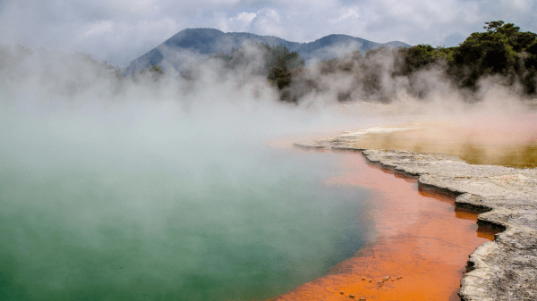 A body of water covered in steam with mountains and sky in the background; the shore to the right has orange outlining it
