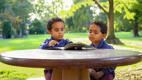 Two children sit at a wooden table with a book in front of them and a grassy field and trees behind them