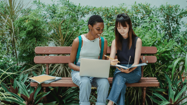 Two people sit on a park bench with notebooks to the left of them; the person on the left has a laptop in their lap, the person on the right is holding a notebook and pencil; behind them is an assortment of green foliage