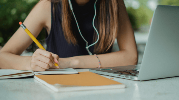 A person, head cut off by the image ending at the top, sits at a table outside with a laptop on it; they're wearing earbuds and holding a pencil over some paper and a notebook