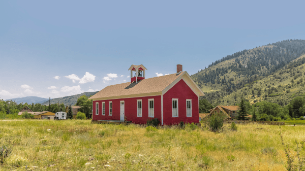 A red schoolhouse sits in the center of a green grassy field, behind it is a green tree-dotted hill and a slightly cloudy blue sky