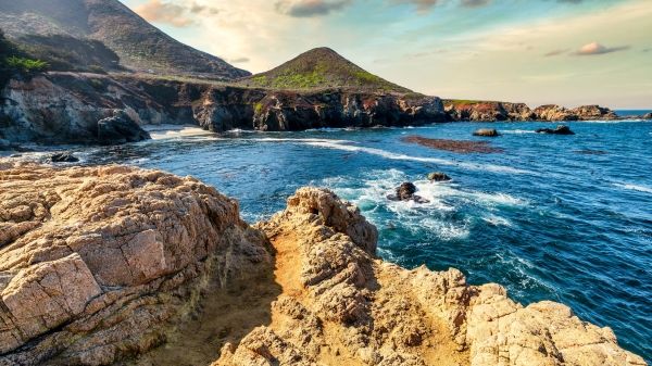 A rocky outcropping lets out onto the ocean; behind it is a green hill and cloudy sky