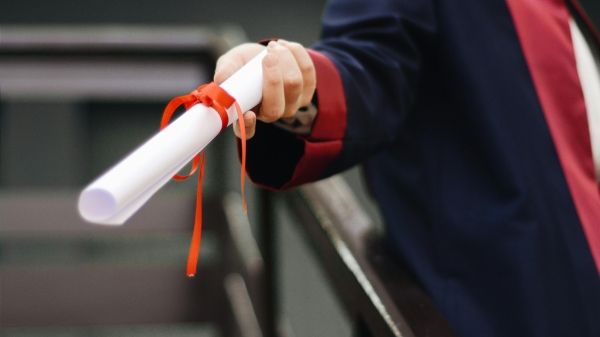 A hand holds out a rolled piece of paper tied with an orange ribbon; the person is wearing a graduation robe