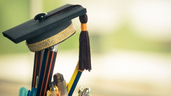 A graduate cap sits on top of pencils in a cup in front of a blurred greenish background
