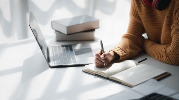 Person writing in a notebook with laptop and books to the side