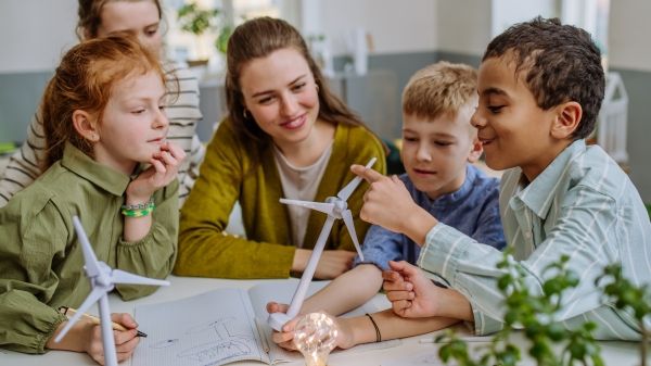 Two pairs of students sit on either side of an instructor; the furthest right student is holding a scale wind turbine; papers and another wind turbine sit on the table in front of them