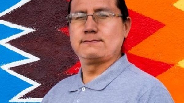 Profile photo of a man wearing glasses, hair pulled back, and staring straight at the camera. He stands in front of a colorful, geometric backdrop