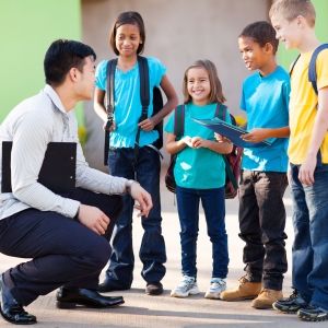 Teacher crouching next to four elementary aged students who are smiling and wearing backpacks.