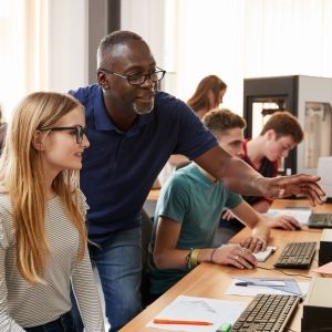 Teenage students working at row of computers. Teacher appears to be helping one student, as they look at the monitor together. 