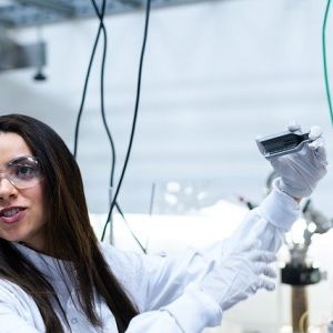 A woman wearing safety glasses in a lab setting with wires in the background, holding a metallic bracket