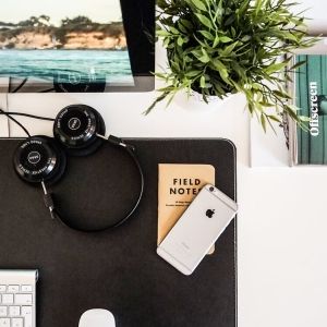 Photo of a desk with a keyboard, mouse, phone, notebook, headphones, computer screen, plant, and book on top