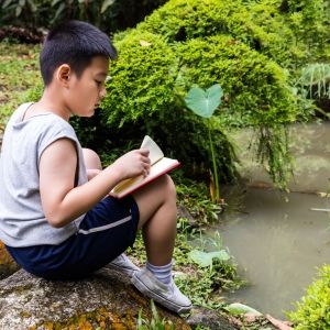Child sitting on a large rock along a pond, reading a book.