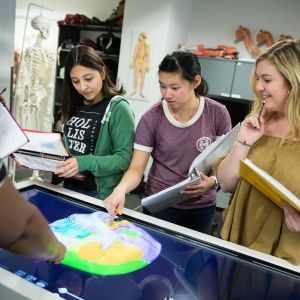 Four young women with open textbooks, point at a display screen