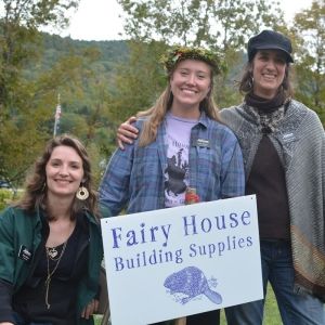 Three light-skinned and long-haired people smile behind a yard sign that reads "Fair House Building Supplies." The person to the left crouches, and the two people to the right stand. Behind them, green grass and trees in late summer foliage.
